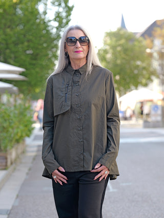 Frau trägt Rundholz Bluse Jungle Print mit Kopfleiste und Brusttasche. Die Bluse hat eine A-Form mit Falten. Sie kombiniert sie mit Sonnenbrille und hält die Hände in den Taschen.