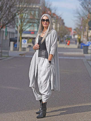 Frau trägt das Rundholz Kleid 3200906 grau als Mantel, kombiniert mit schwarzen Stiefeln und Sonnenbrille, auf einer Straße. Das Kleid hat eine durchgehende Knopfleiste und raffinierte Kellerfalten.