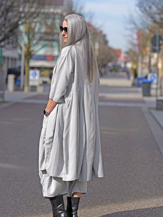 Frau trägt das Rundholz Kleid 3200906 grey, ein vielseitiges Kleidungsstück, als langen Mantel mit durchgehender Knopfleiste und stilvollen Kellerfalten, kombiniert mit schwarzen Stiefeln.