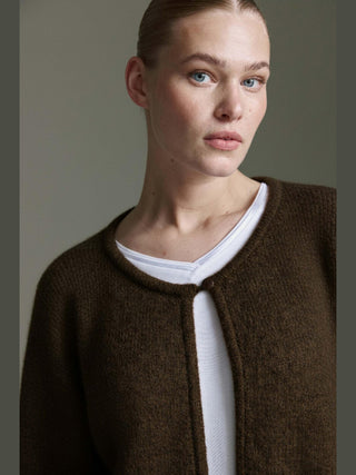 Nahaufnahme eines jungen Erwachsenen mit der Cotton Candy Ona Cardigan espresso, einem dunkelbraunen Strickcardigan mit tiefem Rundhalsausschnitt und einem Knopfverschluss, über einem weißen T-Shirt getragen.