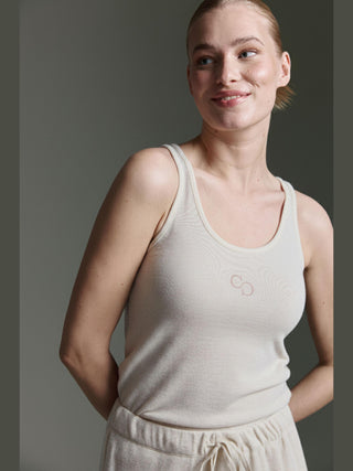 Frau trägt das Cotton Candy Roca Top offwhite, ein ärmelloses, geripptes Tanktop mit dezentem Logo auf der Brust, kombiniert mit passenden Hosen, ihre Haare sind zurückgebunden, sie lächelt sanft.