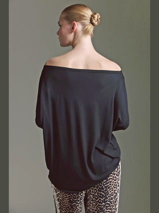 Person von hinten in einem lockeren, schulterfreien Cotton Candy Ondra Tee Schwarz, das elegant über den Oberkörper fällt, kombiniert mit auffälligen Leopardenhose. Silhouette und Kontrast stehen im Mittelpunkt.