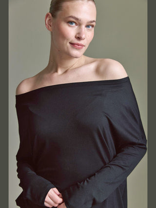 Person trägt das Cotton Candy Ondra Tee Black, ein langärmeliges, schulterfreies Oberteil, das elegant die Schultern betont. Der lockere Schnitt bietet vielseitige Styling-Möglichkeiten für legere oder elegante Outfits.