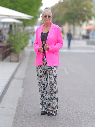 Frau trägt den Emily Damenblazer neon pink, einen lässigen Blusen-Blazer mit Krempelärmeln und aufgesetzten Patten aus gewaschener Leinenqualität, kombiniert mit gemusterten schwarz-weißen Hosen.