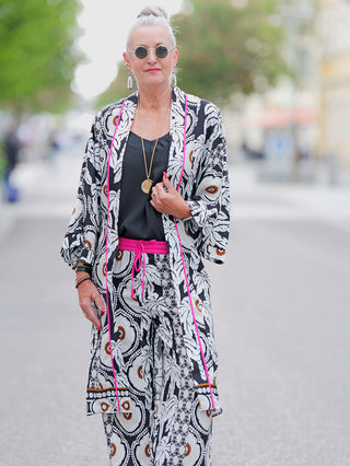 Emily Kimono pink/schwarz/sand aus fließender Viskose, lässig gebunden mit Gürtel, zeigt ein Ethno-Print-Muster. Die Frau trägt Sonnenbrillen und Schmuck, was den eleganten Look ergänzt.