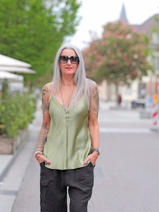 Person trägt das OFF#DLY Top Button green: ärmelloses, leichtes Viskose-Top mit Knopfleiste, kombiniert mit locker sitzenden Cargo-Hosen. Entspannte, urbane Umgebung deutet auf sommerlichen Straßenstil hin.