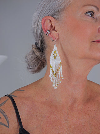 Nahaufnahme eines Kopfes mit grauen Haaren im Dutt, zeigt OFF#DLY BoHo Ohrschmuck white/gold: lange, fransige Ohrringe aus weißen und goldenen Perlen, ergänzt durch kleine silberne Ringe und Manschetten.