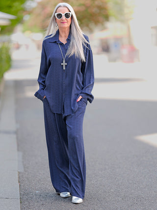 Frau mit grauem Haar trägt die OFF#DLY Summer Shirt Teresa blue, eine Longbluse mit Knopfleiste. Sie trägt eine passende blaue Hose und weiße Sonnenbrille.