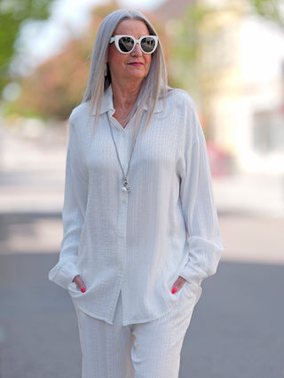Frau in weißem OFF#DLY Summer Shirt Teresa mit langen Ärmeln und Knopfleiste, trägt Sonnenbrille. Shirt aus 58% Rayon und 42% Viskose.