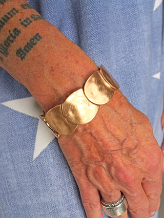 Nahaufnahme eines Handgelenks mit einem OFF#DLY Armband gold matt. Das breite, metallische Armband präsentiert sich in einer eleganten, matten Optik und ist auf einem Handgelenk mit Tätowierung abgebildet.