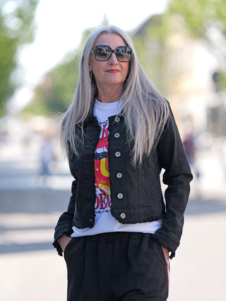 Frau mit langen grauen Haaren trägt die OFF#DLY Colordenim Jacket schwarz mit ausgefransten Kanten, während sie eine Sonnenbrille trägt. Die Jacke hat ein klassisches Jeansjacken-Design.