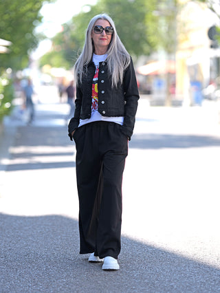 Eine Frau mit langen grauen Haaren trägt die OFF#DLY Denim Jacket black, eine klassische Kurzjacke im Jeansjacken-Design mit ausgefransten Kanten. Sie kombiniert sie mit einer schwarzen Hose.
