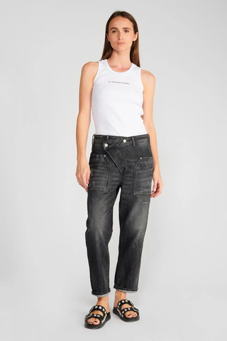 Jean Femme Cosy Pocket, getragen von einer Frau in einem weißen Tanktop; schwarze Jeans mit einzigartigem Cross-over-Verschluss und lockerer Passform, kombiniert mit schwarzen Sandalen mit silbernen Nieten.