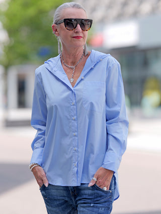 Emily Damenbluse lightblue, getragen von einer Frau mit Sonnenbrille. Die Bluse hat eine Kapuze, aufgesetzte Tasche und lange Ärmel mit Manschette. Baumwollstretch für lässigen Stil.
