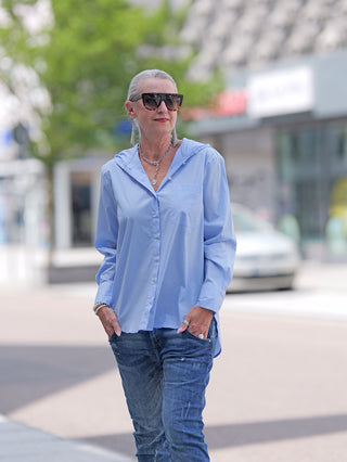 Emily Damenbluse lightblue: Frau trägt hellblaue Bluse mit Kapuze, aufgesetzter Tasche und langen Ärmeln. Kombiniert mit Jeans und Sonnenbrille, lässiger Look. Material: Baumwollstretch.