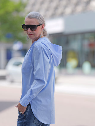 Emily Damenbluse lightblue: Frau trägt eine hellblaue Bluse mit Kapuze, aufgesetzter Tasche und langen Ärmeln mit Manschetten, kombiniert mit lässiger Jeans. Perfekt für einen entspannten, modischen Auftritt.