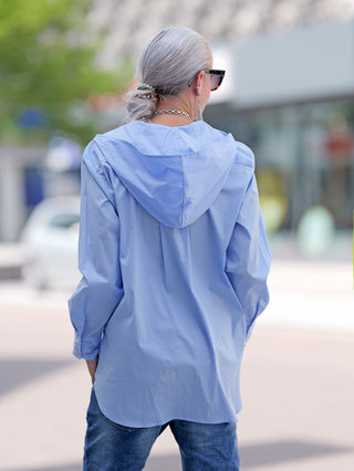 Emily Damenbluse lightblue, getragen von einer Frau. Die Bluse hat eine Kapuze, eine aufgesetzte Tasche und lange Ärmel mit Manschette. Hergestellt aus lässig gewaschener Baumwollstretchware.