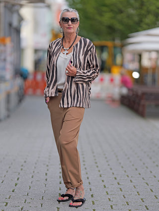 Frau trägt die OFF#DLY Blazer Zebra mit asymmetrischem Schnitt und einem Knopf, kombiniert mit braunen Hosen und schwarzen Sandalen, während sie auf dem Gehweg steht.
