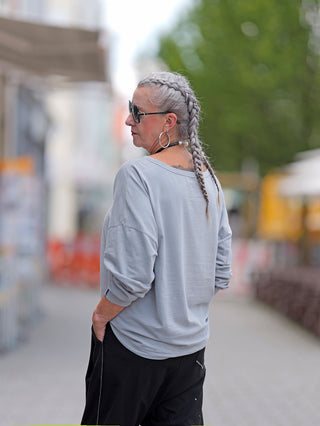 Frau mit geflochtenem Haar und Sonnenbrille trägt ein True Religion Wide Crew Tee Reflective. Das T-Shirt hat einen weiten Rundhalsausschnitt und eine reflektierende Oberfläche.