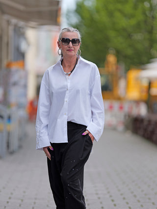 Frau trägt True Religion Bluse Double Cuff Oversized mit großem Kragen und Doppelmanschette, kombiniert mit Sonnenbrille und großen Ohrringen. Bluse aus Baumwolle, lässig und stilvoll.
