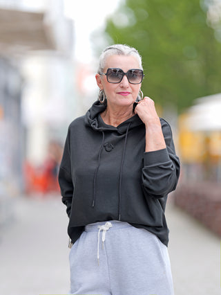 Frau trägt True Religion Hoody schwarz mit Kapuze und Sonnenbrille, aus komfortablem Materialmix. Nahaufnahme zeigt Reißverschluss und modischen Straßenstil, ideal für lässige Anlässe.