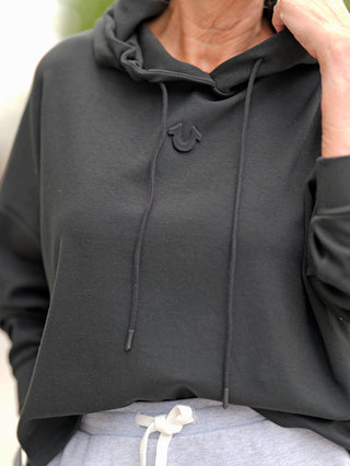 True Religion Hoody schwarz, getragen von einer Person, zeigt die sportliche Passform und den bequemen Materialmix. Markantes Detail: weiße Kordel an der Kapuze, ideal für lässigen, modischen Stil.