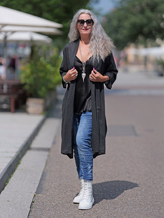 Frau trägt den OFF#DLY Kimono Roxy schwarz auf der Straße, kombiniert mit blauen Jeans und weißen Stiefeln. Der leichte Baumwollkimono ist locker geschnitten und passt bis Größe 42.