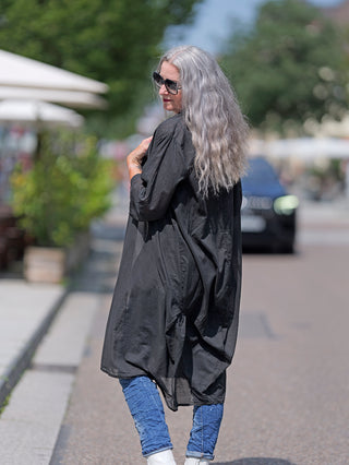 Frau trägt den OFF#DLY Kimono Roxy schwarz, aus leichter Baumwolle, während sie auf der Straße läuft. Kombiniert mit blauer Jeans und weißen Stiefeln, betont der Kimono ihren modernen Stil.