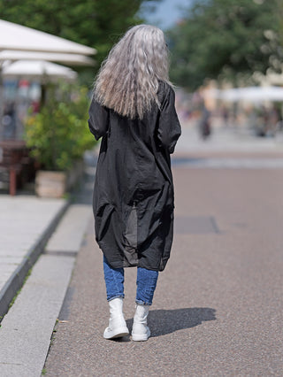 Eine Frau trägt den OFF#DLY Kimono Roxy schwarz, während sie auf der Straße geht, kombiniert mit weißen Stiefeln. Der Kimono ist leicht und aus 100% Baumwolle.