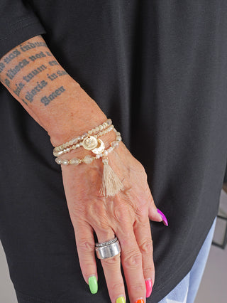 Nahaufnahme eines Arms mit Tattoos, der das OFF#DLY 3 Strang Armband beige trägt. Das elastische Armband aus Metall und Glasperlen hat eine Länge von 18 cm und ist als One Size erhältlich.