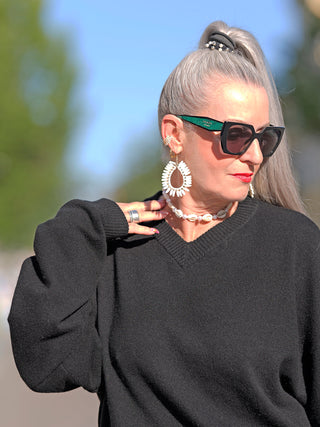 Eine Frau trägt die Gitta Banko Juno black Sonnenbrille und einen schwarzen Pullover, mit einem weißen Ohrring im Nahbereich sichtbar.