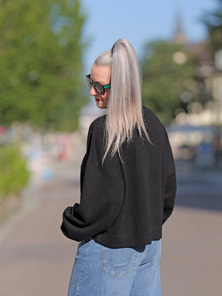 Frau mit langen Haaren trägt die Gitta Banko Juno black Sonnenbrille und einen schwarzen Pullover; modisches Straßenoutfit mit Jeans, ideal für lässige Outdoor-Anlässe.