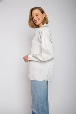 Emily Bluse 102 offwhite: Eine Frau trägt eine oversized Hemdbluse mit überschnittener Schulter und Knopfblende, gefertigt aus leicht fließendem Viskose Satin. Die Bluse wirkt lässig durch eine Rückenteilfalte.