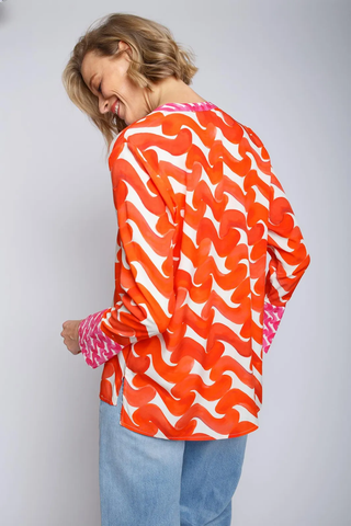Emily Bluse 490 dark/orange/pink: Frau trägt eine farbenfrohe Bluse mit kleinem Kragen, V-Ausschnitt und 1/1 Ärmeln, kombiniert mit blauen Jeans, lächelnd über die Schulter blickend.