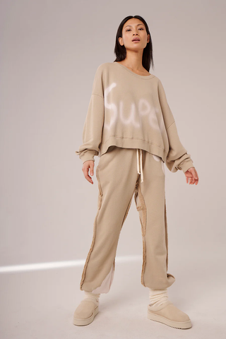 Li Bergen Sweater Graz bleached: Frau trägt lässigen Camel-Pullover mit verwaschenem Super-Aufdruck, kombiniert mit Hose. Der Pullover hat lange Ärmel und ist aus Baumwolle.