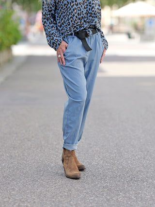 Person trägt OFF#DLY Jogger Antonia hellblau mit lockerem Schnitt und Gürtel, kombiniert mit blauen Jeans und braunen Stiefeln. Hände in den Taschen, keine Gesäßtaschen sichtbar.