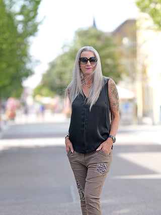 Frau mit Sonnenbrille trägt OFF#DLY Top Tanja, ein ärmelloses schwarzes Seidentop mit breiten Trägern und V-Ausschnitt, auf der Straße. Sichtbar sind auch Tattoos auf ihrem Arm.