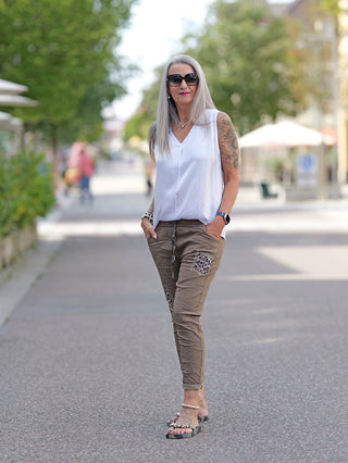 Frau auf dem Gehweg trägt das OFF#DLY Top Tanja white mit breiten Trägern und V-Ausschnitt. Die Arme zeigen Tattoos, die langen Haare sind offen, und sie trägt Sonnenbrillen.