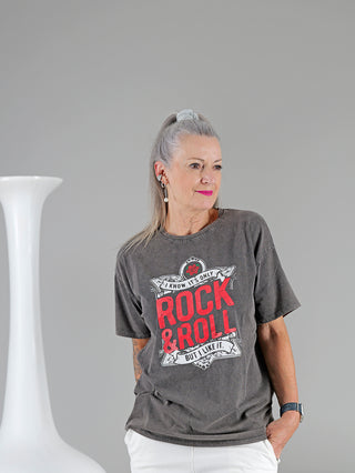 Frau in grauem OFF#DLY Tee ROCK mit rotem Aufdruck, stehend neben einer weißen Vase. T-Shirt aus 95% Baumwolle, 5% Elastan, Einheitsgröße bis Größe 40.