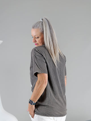 Frau trägt das OFF#DLY Tee ROCK, ein graues Rundhalt-T-Shirt mit Frontprint. Ein modisches Accessoire, eine Uhr, ist am Handgelenk zu sehen. Materialmix aus Baumwolle und Elastan.