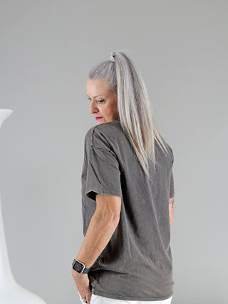 Frau mit grauen Haaren trägt das OFF#DLY Tee ROCK, ein graues Rundhals-T-Shirt mit Frontprint. Ihr Arm zeigt eine Uhr und einen Perlenring. Material: 95% Baumwolle, 5% Elastan.