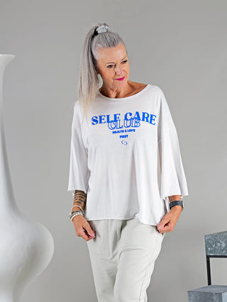 Frau mit grauem Haar trägt das Cotton Candy Tee Helin mit 3/4 Ärmeln und SELF CARE CLUB Frontprint. Das Shirt wirkt lässig und bequem.