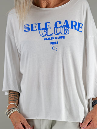 Frau trägt das Cotton Candy Tee Helin mit 3/4 Ärmeln und Rundhalsausschnitt. Aufdruck SELF CARE CLUB sichtbar. Bracelet und Tattoo am Arm ergänzen den Look.