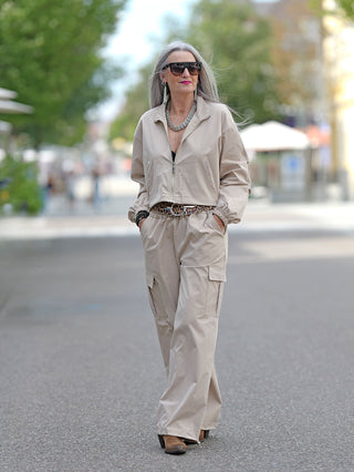 Frau trägt den OFF#DLY Anzug Tina beige mit Gummizug an Bund und Beinabschlüssen, seitlichen Cargotaschen und einer Jacke mit Zipper, dazu große Sonnenbrille.