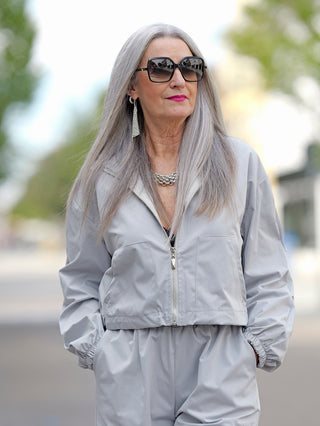 Eine Frau trägt den OFF#DLY Anzug Tina grey mit Sonnenbrille, bestehend aus einer Jacke mit Bund und Hose mit Gummizug und Cargotaschen, ergänzt durch modische Accessoires.