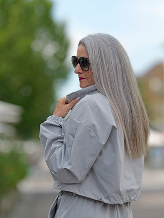 Frau mit langen grauen Haaren und Sonnenbrille trägt den OFF#DLY Anzug Tina grey, bestehend aus einer Jacke mit Gummizug und einer Hose mit seitlichen Cargotaschen und Gummizug am Bund.