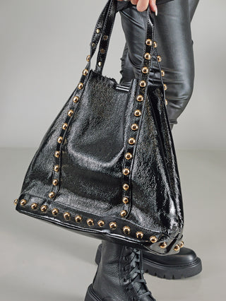 OFF#DLY Lina Shopper black aus glänzendem Fake-Leder mit goldfarbenen Nieten, getragen von einer Person zu schwarzen Lederhosen. Der Shopper hat ein Teddy-Innenfutter und eine kleine Innentasche.