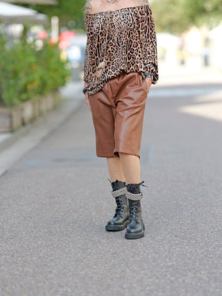 Person trägt die OFF#DLY Short Lilly, eine stilvolle Fake-Leder-Short mit weitem Bein, zusammen mit schwarzen Stiefeln. Das Modell zeigt eine lässige, modische Outdoor-Kombination.