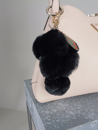 OFF#DLY Taschenanhänger black, ein schwarzer, plüschiger Taschenanhänger aus Polyester, mit einem auffälligen goldenen Ring und einer grünen Applikation auf der Vorderseite. Größe: 15 x 10 cm.