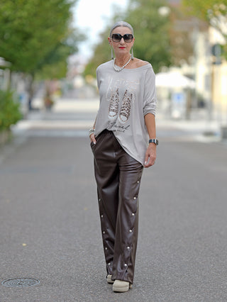Frau trägt OFF#DLY Doreen Pant, eine lässige Wide Leg Fake Lederhose mit seitlichen Druckknöpfen und Gummizug am Bund. Sie kombiniert die Hose mit einem grauen Shirt und Sonnenbrille.